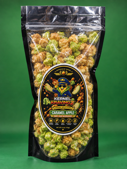 A bag of Kernel Kravings Caramel Apple popcorn on a solid green background. The product is packaged in a clear, resealable black stand-up pouch with a prominent circular label showing a cartoon character and the flavor name.